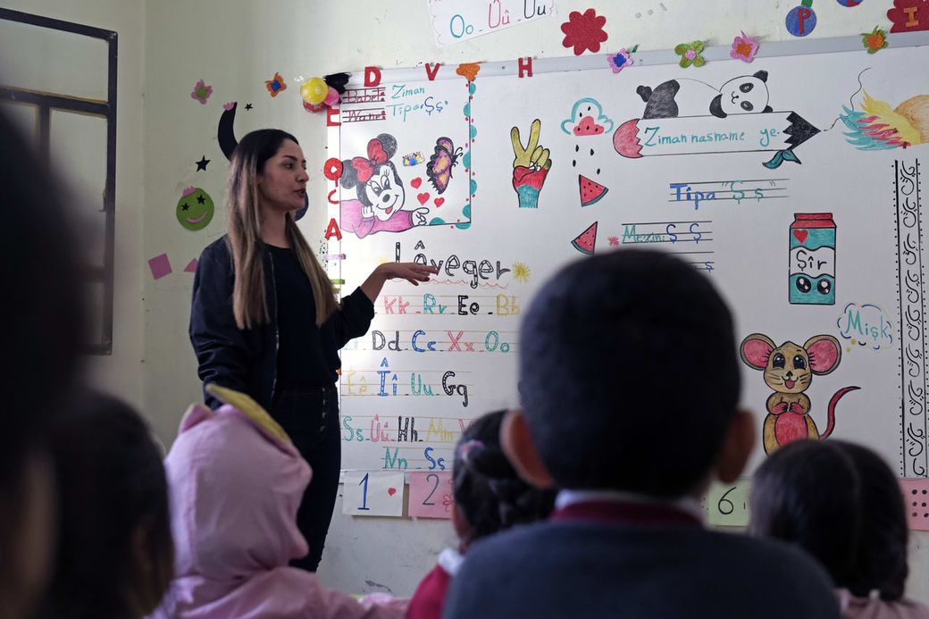 A teacher points to a colorful alphabet chart on the wall as young students watch attentively in a classroom setting.