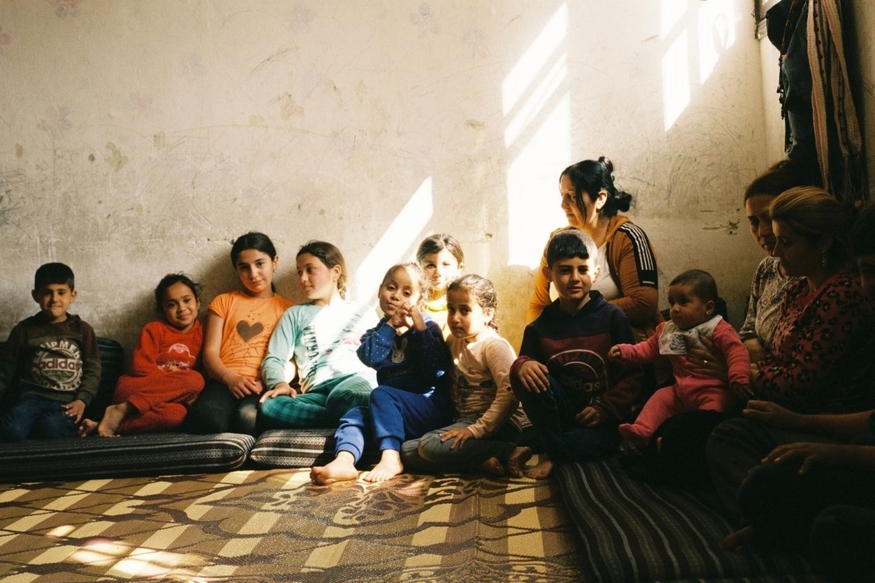 A group of children and women sitting on mats in a sunlit room, with light streaming in from a window, creating a warm, communal atmosphere.