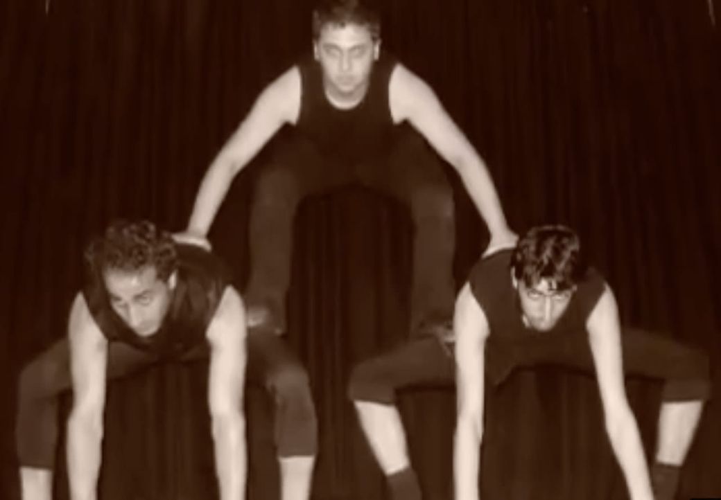 Three individuals perform a human pyramid, with two crouched below and one balancing on top, against a dark curtain background.