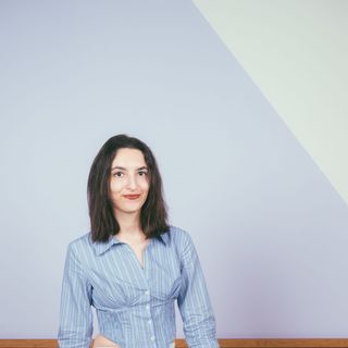 A woman in a striped blue shirt and light jeans sits on a wooden stool against a light purple background with a wooden panel.