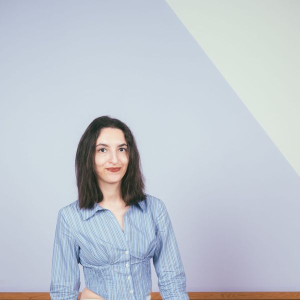 A woman in a striped blue shirt and light jeans sits on a wooden stool against a light purple background with a wooden panel.