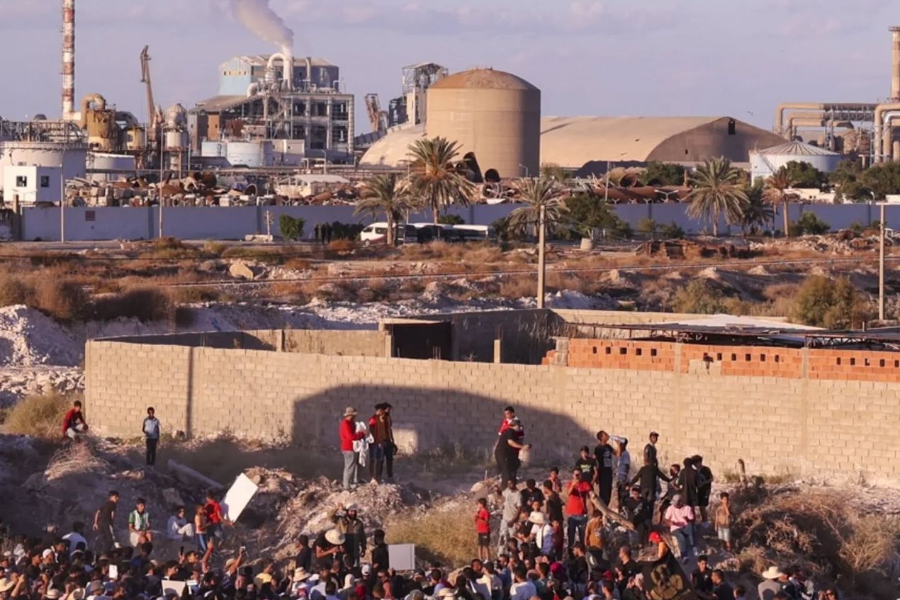 Op de voorgrond: een manifestatie tegen de fosforfabriek in Gabès, Tunesië. Op de achtergrond de fabriek zelf, met een duidelijk zichtbare rookwolk.
