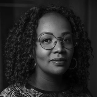 A person with curly hair wearing glasses and colorful attire looks directly at the camera with a calm expression.