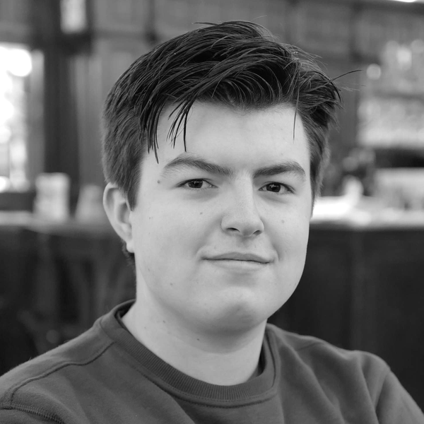 A person with short dark hair wearing a red sweater is smiling in a cozy, warmly-lit room with wooden decor.