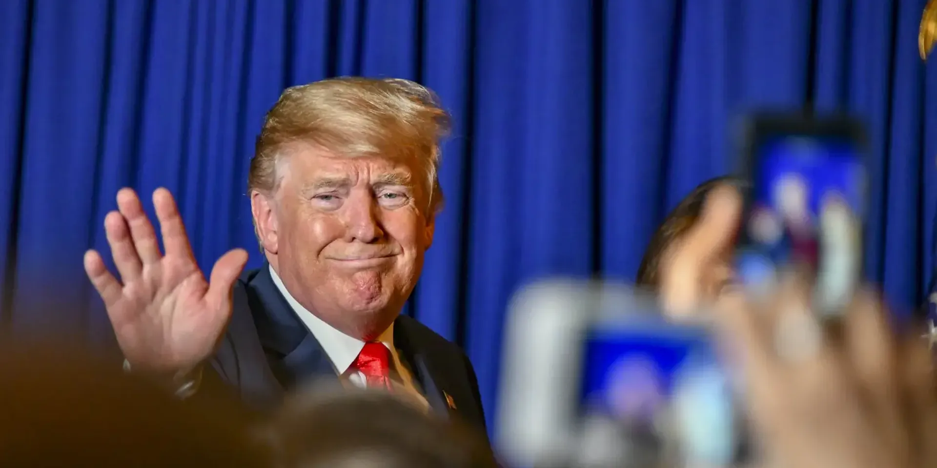 president Trump voor een blauw gordijn, zwaaiend naar aanwezigen in de zaal