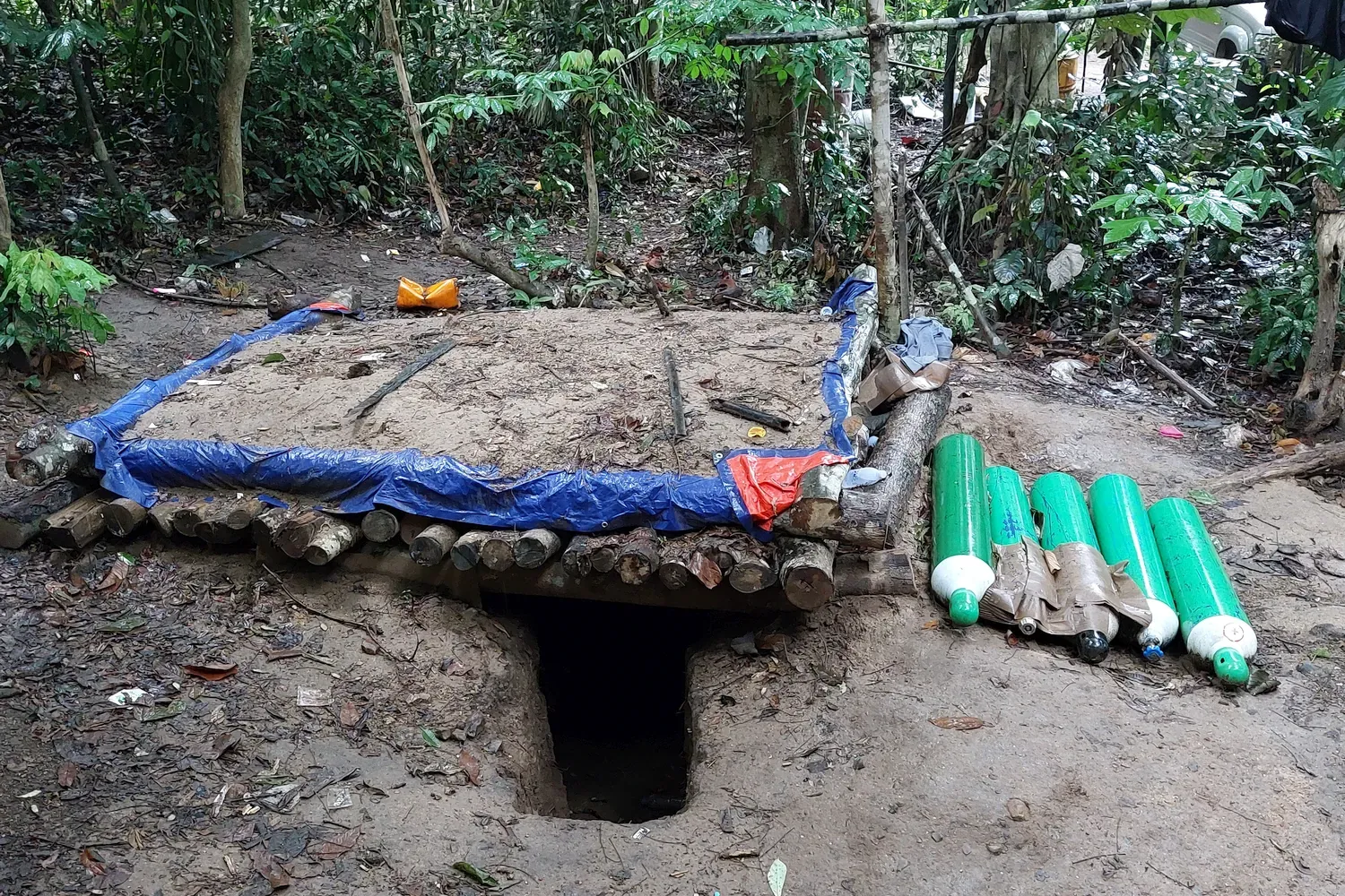 Een met boomstammen en zand bedekte schuilplaats tegen bombardementen. Naast de schuilplaats liggen enkele cilinders op de grond.