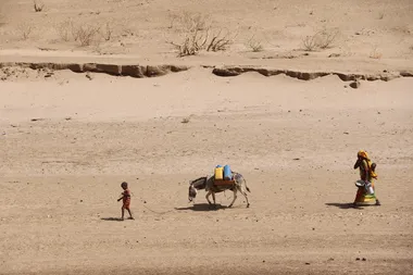 een jongen loopt voor een met waterbidons bepakte ezel in de woestijn, gevolgd door een vrouw.