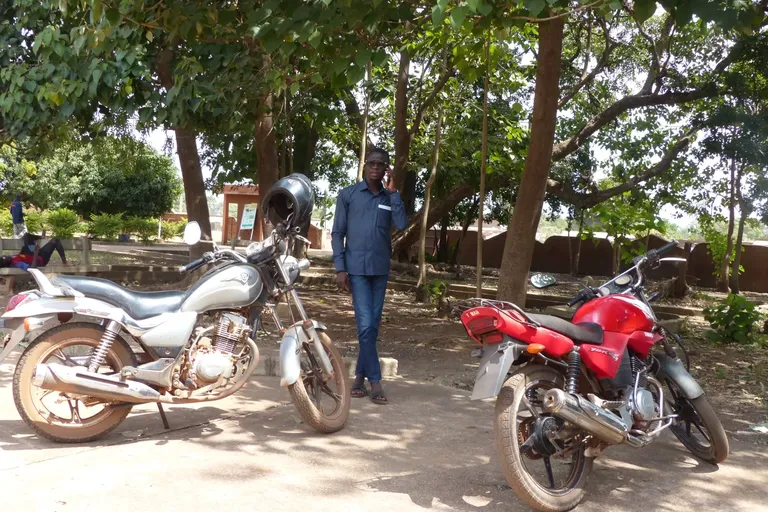 Een man staat in de schaduw van enkele grote bomen tussen twee brommers