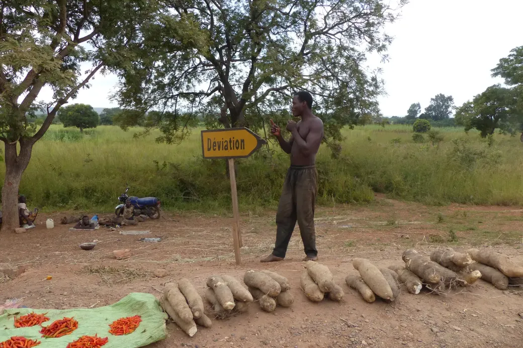 Een man staat naast een lading maniokwortels en het verkeersbord ‘Déviation’