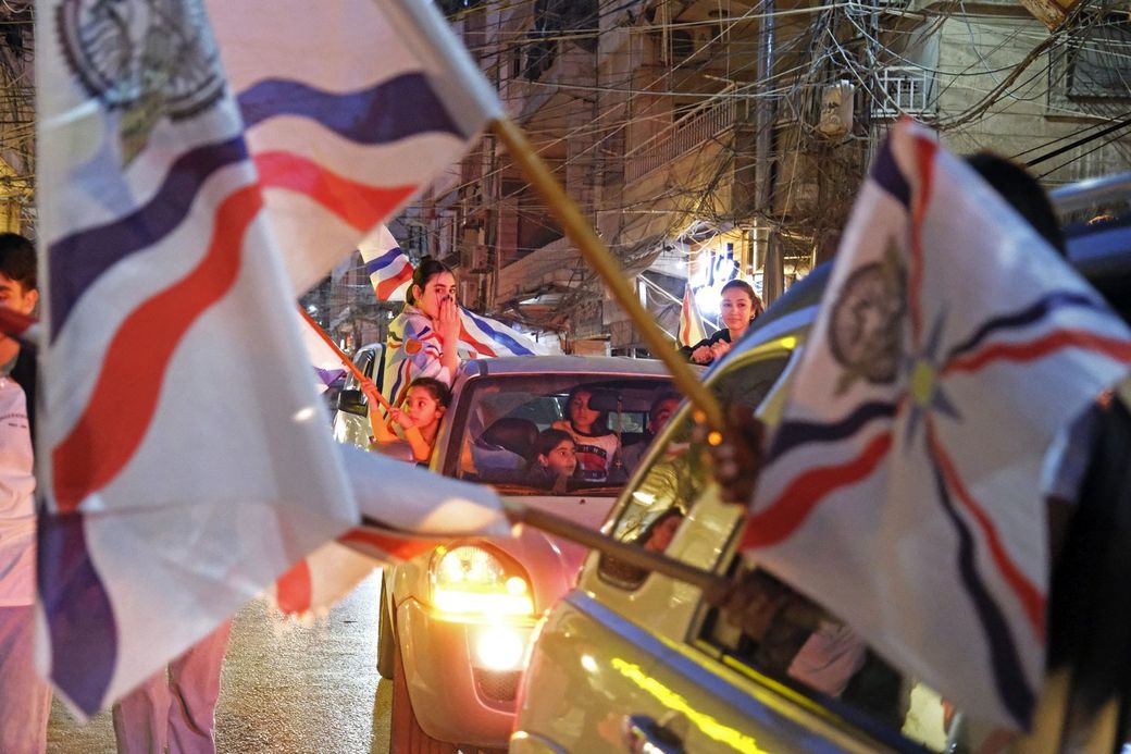 People in cars celebrate with flags on a busy street at night, with buildings and wires visible in the background.