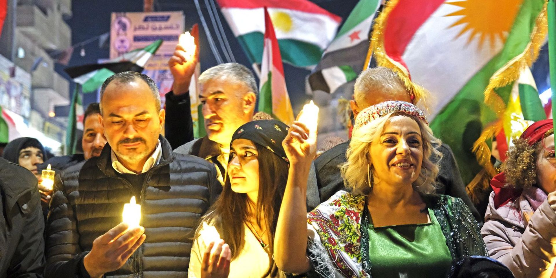 Crowd in colorful attire holding candles, with vibrant flags in the background during a nighttime gathering.