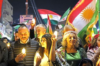 Crowd in colorful attire holding candles, with vibrant flags in the background during a nighttime gathering.