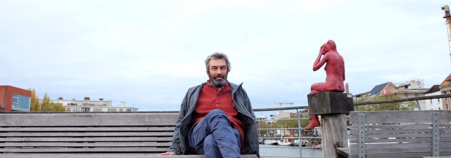John Vandaele zit op een bank aan de waterkant naast een rood standbeeld