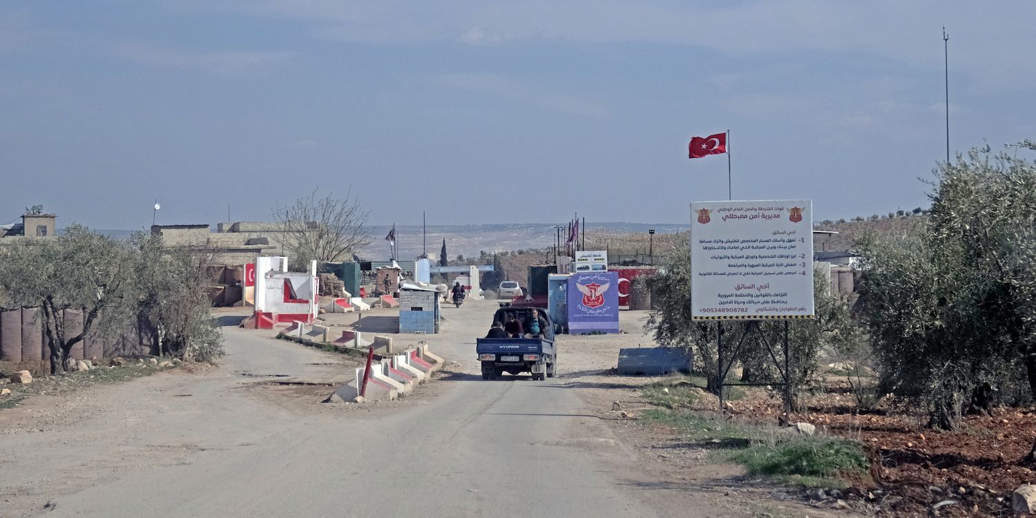 A rural road leads to a checkpoint with a Turkish flag. A truck approaches barriers, surrounded by trees and signs with Arabic text.