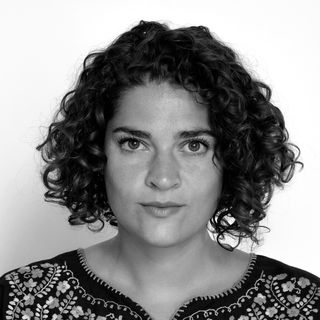 A woman with curly brown hair wearing a black top with intricate yellow floral embroidery, set against a plain white background.
