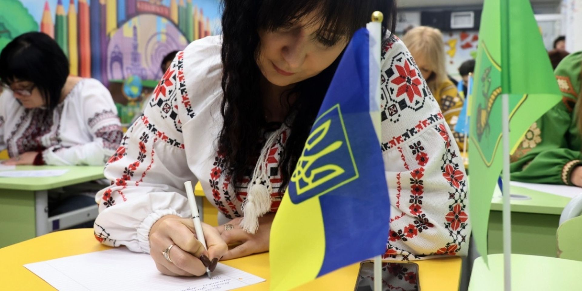 Woman in embroidered blouse writes at a desk with a small Ukrainian flag, surrounded by others in traditional attire.