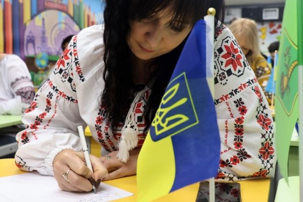 Woman in embroidered blouse writes at a desk with a small Ukrainian flag, surrounded by others in traditional attire.