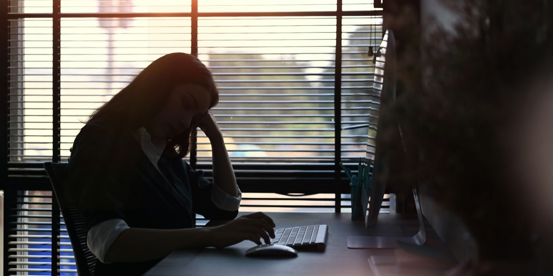Silhouetted woman in office working on a computer, with sunlight streaming through blinds.