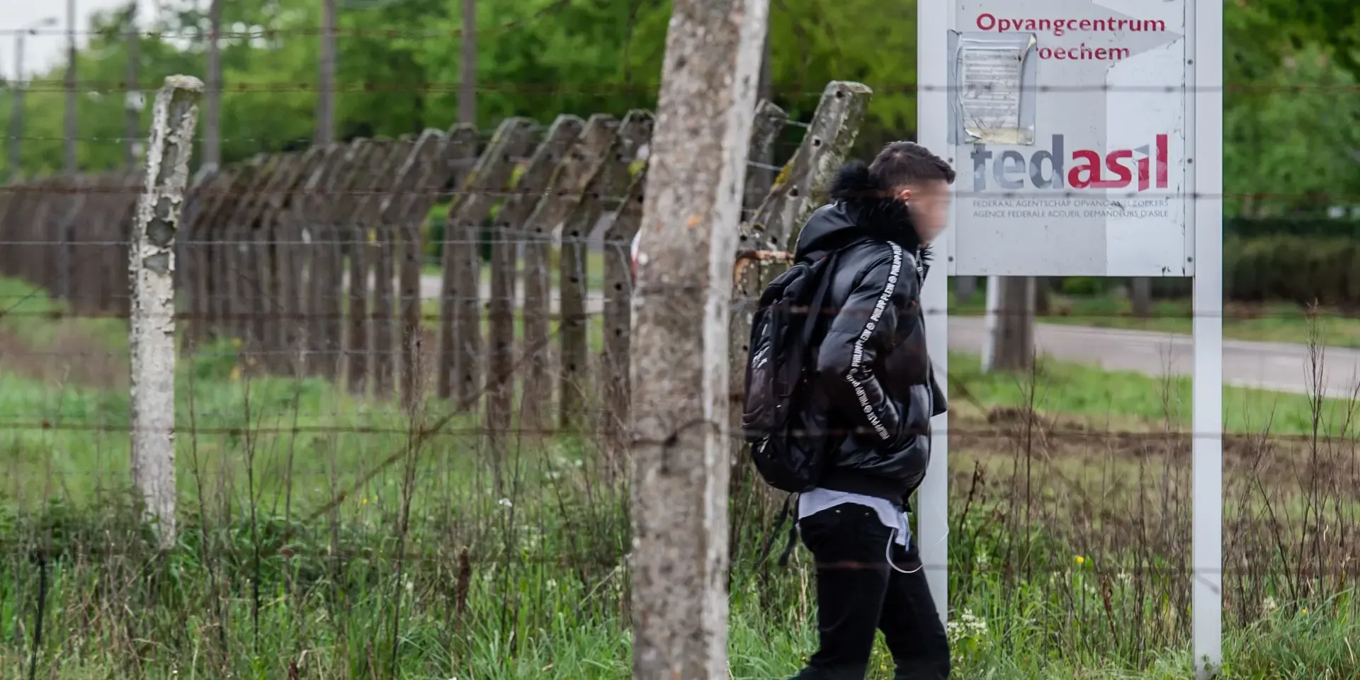 Een man verlaat een met prokkeldraad omgeven terrein en wandelt voobij een bord waarop de vermelding 'Opvangcentrum Broechem, en fedasil staat.