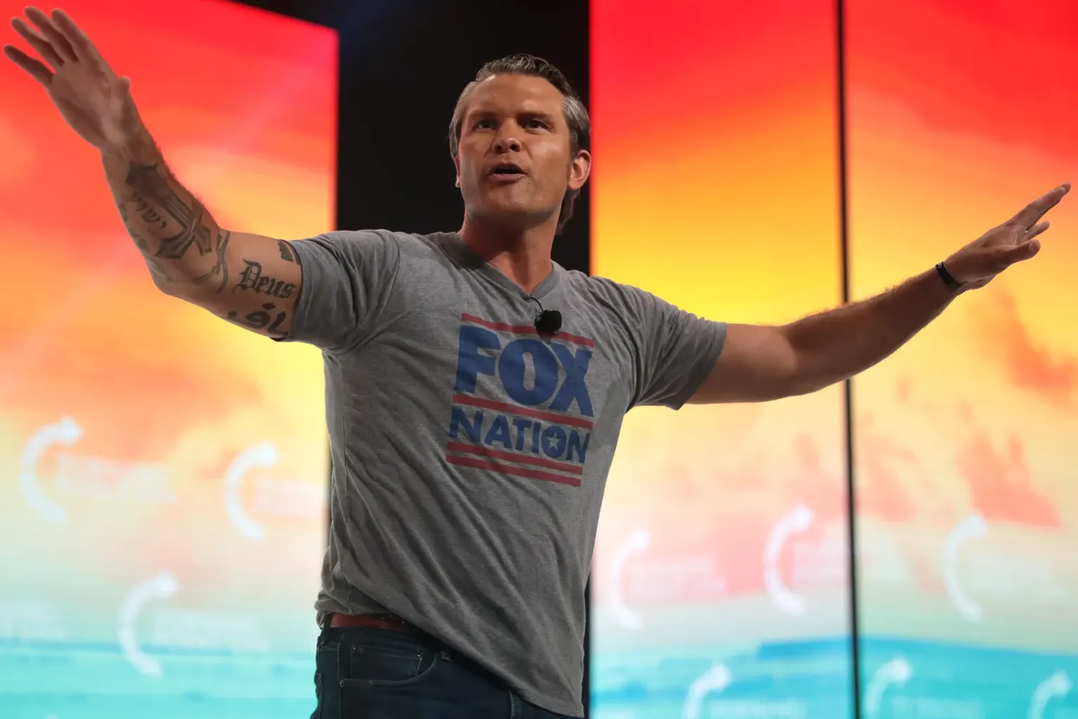 De Amerikaanse minister van oorlog Pete Hegseth staat met gespreide armen op een podium