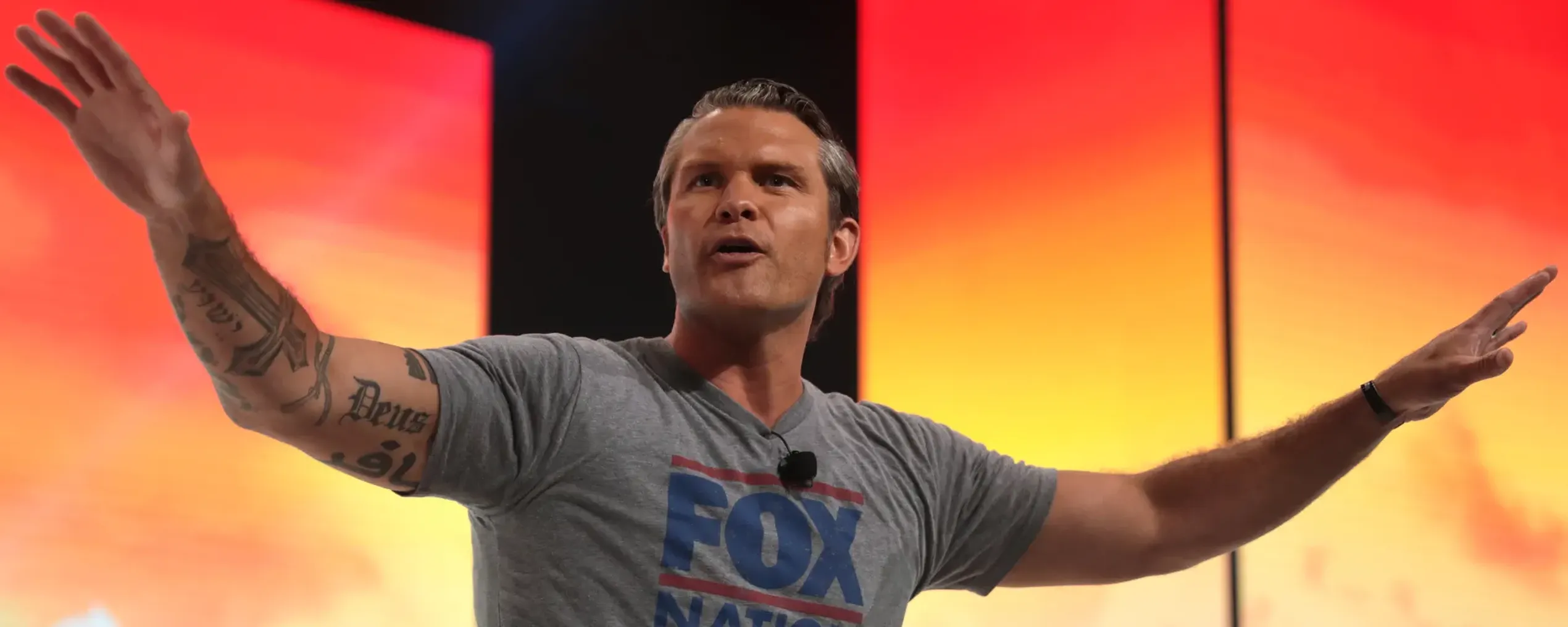 De Amerikaanse minister van oorlog Pete Hegseth staat met gespreide armen op een podium