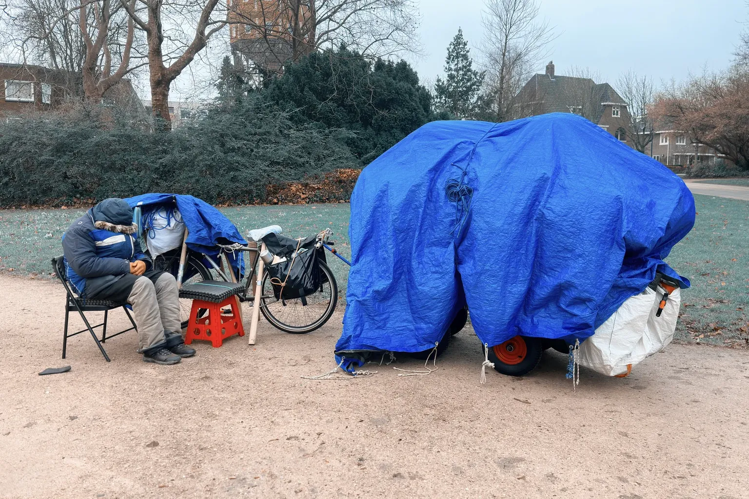 Een dakloos persoon zit dik ingeduffeld op een vouwstoel nast een kar met zijn schamele bezittingen
