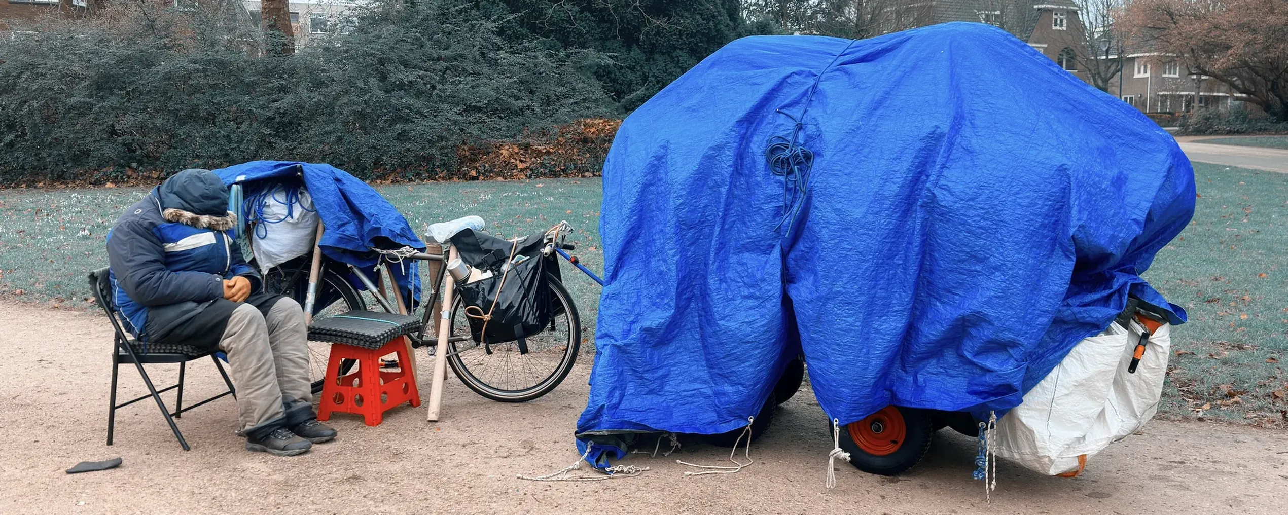Een dakloos persoon zit dik ingeduffeld op een vouwstoel nast een kar met zijn schamele bezittingen