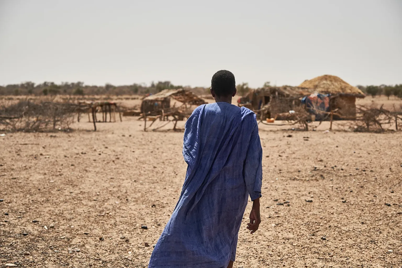 Een man in blauwe klederdacht staat met zijn rug naar de camera. Op de achtergrond silhouetten van een klein dorpje in Mauritanië