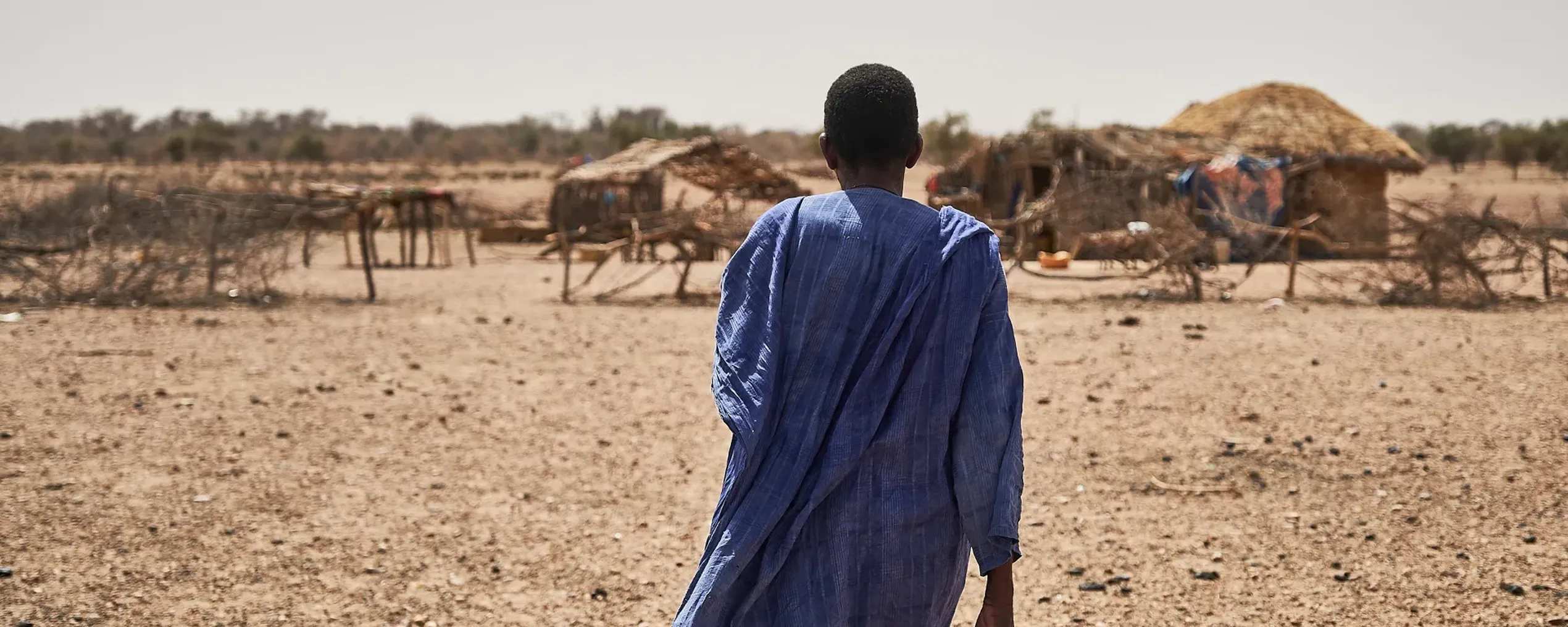 Een man in blauwe klederdacht staat met zijn rug naar de camera. Op de achtergrond silhouetten van een klein dorpje in Mauritanië