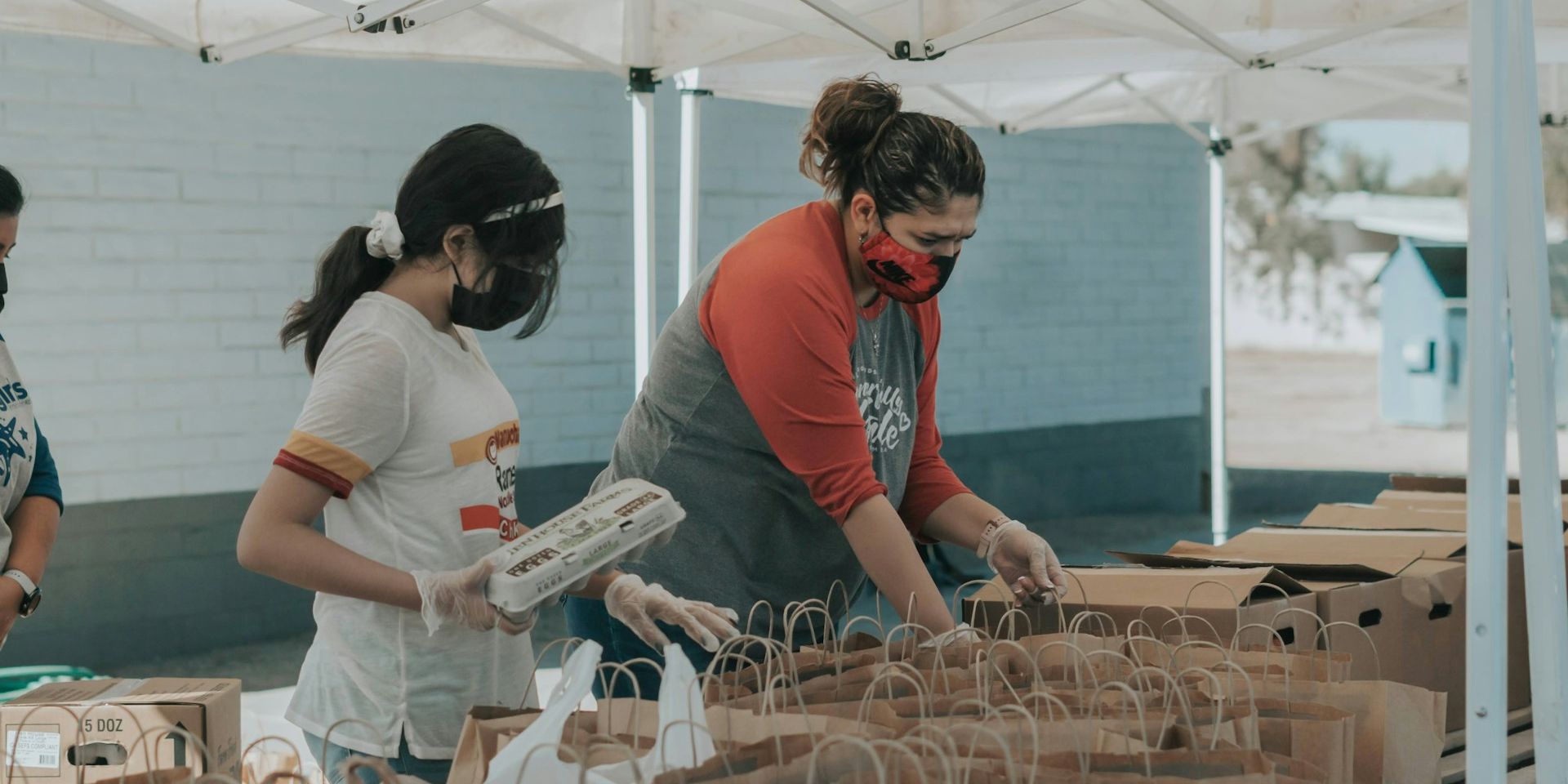 People wearing masks and gloves organize food packages under a tent, with rows of paper bags on tables.