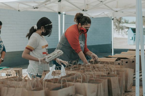 People wearing masks and gloves organize food packages under a tent, with rows of paper bags on tables.