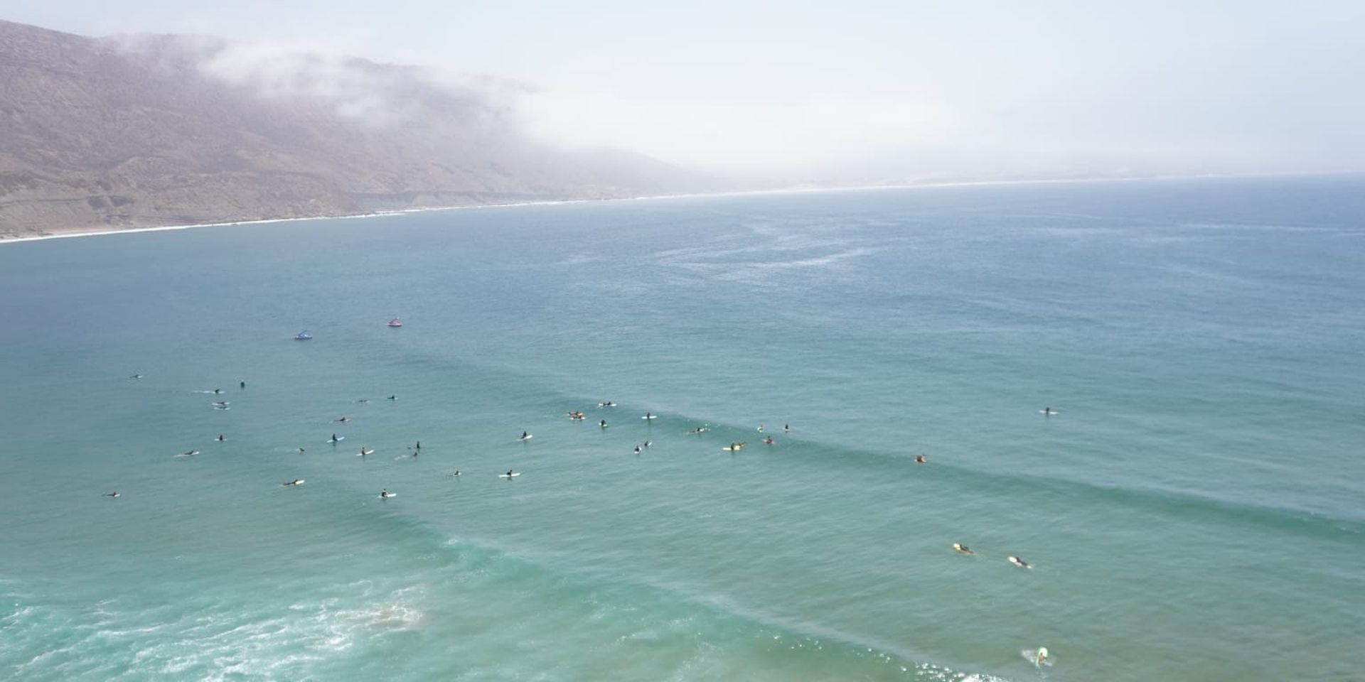 Surfers on a vast ocean, with mist-covered mountains in the background under a clear sky.