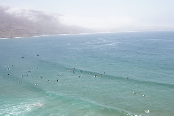 Surfers on a vast ocean, with mist-covered mountains in the background under a clear sky.