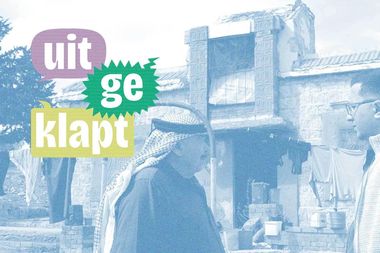 Two men converse in front of a rustic stone building, with colorful speech bubbles displaying the text "uitgeklapt."