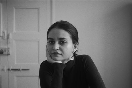 Black and white image of a person with long hair, wearing a turtleneck, sitting at a table, resting their chin on their hand, looking at the camera.
