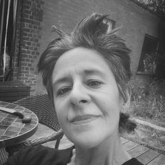 Black and white selfie of a person with short hair smiling outdoors, sitting by a tiled table and brick building.