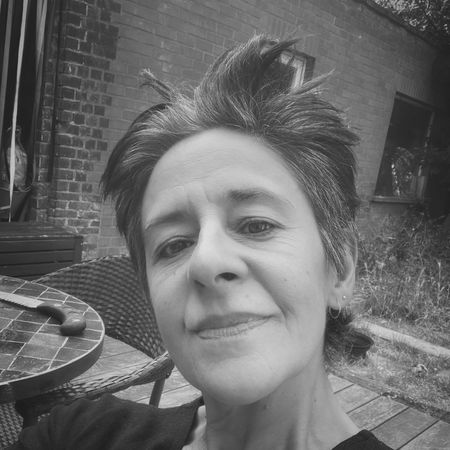 Black and white selfie of a person with short hair smiling outdoors, sitting by a tiled table and brick building.
