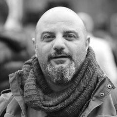 A man with a beard and mustache is wearing a knitted scarf and a green jacket. The background is blurred, indicating a street or outdoor setting.