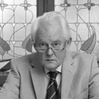 Elderly man with white hair in a beige suit and red tie, sitting indoors with a decorative stained glass window behind him.