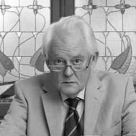 Elderly man with white hair in a beige suit and red tie, sitting indoors with a decorative stained glass window behind him.