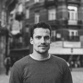 Black and white photo of a person in a sweater, standing outdoors in a cityscape with blurred buildings in the background.