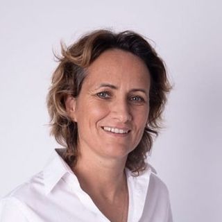 Smiling person with short, wavy hair wearing a white shirt against a plain background.