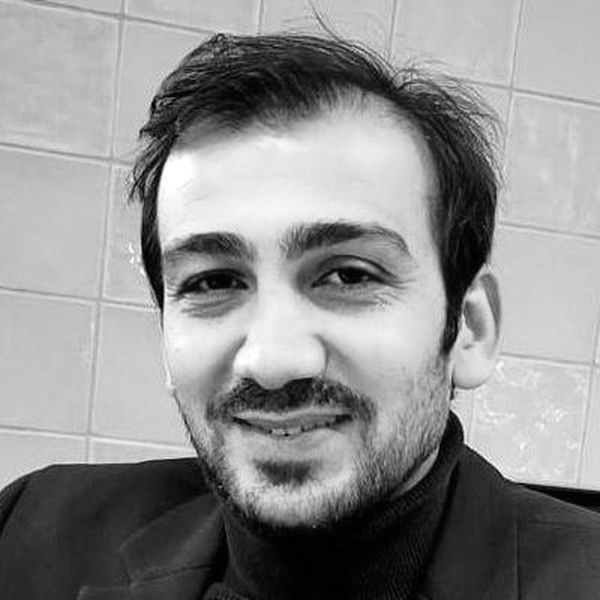 Smiling man with short hair and a beard, wearing a dark turtleneck and blazer, in front of a tiled wall.