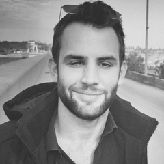 A man with short hair and a beard, wearing sunglasses on his head and a dark jacket, smiles in a grayscale outdoor setting.
