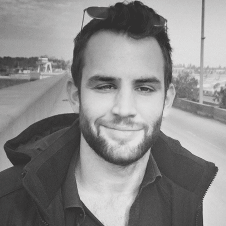 A man with short hair and a beard, wearing sunglasses on his head and a dark jacket, smiles in a grayscale outdoor setting.