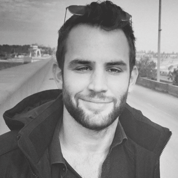 A man with short hair and a beard, wearing sunglasses on his head and a dark jacket, smiles in a grayscale outdoor setting.