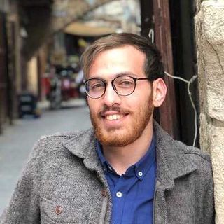 Young man with glasses and a beard smiling, standing in a narrow alley. He wears a blue shirt and gray jacket. Blurred urban background.