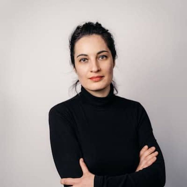 A woman with dark hair in a bun wearing a black turtleneck stands with arms crossed against a plain white background.