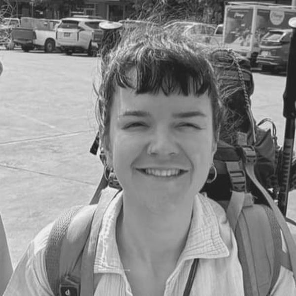 A person smiling outdoors, wearing a backpack and a light-colored shirt. Cars and trees are in the background. Black and white.
