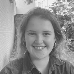 Smiling person with hoop earrings and shoulder-length hair, standing outdoors with trees and a brick wall in the background, in black and white.